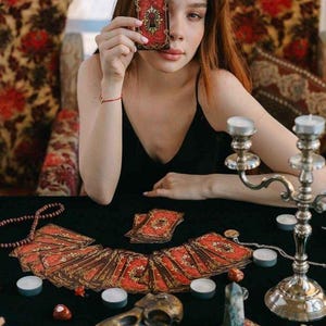May include: A person holding a tarot card with a key design. A spread of tarot cards with red and gold designs is on a black surface. Candles, a bird skull, and crystals are also present.