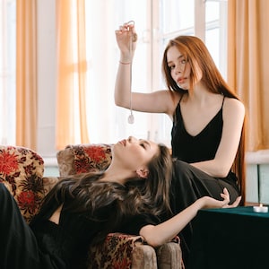 May include: Two women, one holding a pendulum over the other. The woman holding the pendulum wears a black top. The other woman lies on a floral couch, wearing black. A silver pendulum with a clear crystal is suspended. The room has large windows and orange curtains.