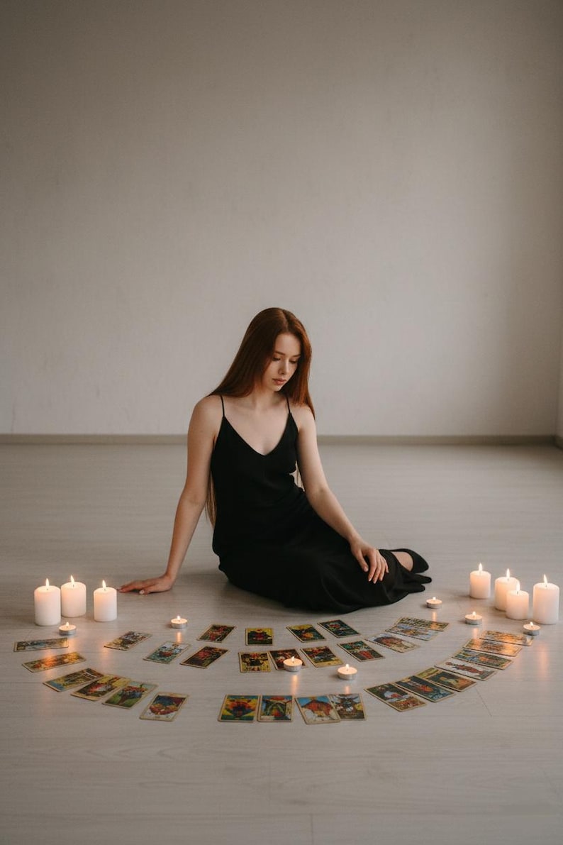 May include: A person in a black dress sits on the floor surrounded by tarot cards and lit candles. The cards are arranged in a semi-circle, with several white candles placed around the perimeter. The scene evokes a mystical or spiritual theme.
