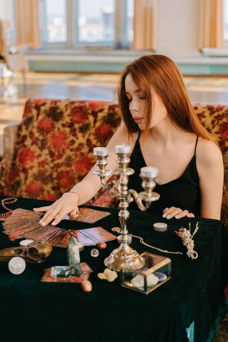 May include: A person in a black top sits at a table covered in a dark green cloth, surrounded by tarot cards, candles, and decorative objects. A silver candelabra with three candle holders is in the foreground. The background features a floral patterned sofa.