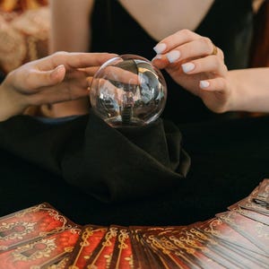 May include: A transparent crystal ball is held between two hands, with tarot cards fanned out below. The cards have red and gold designs. The scene suggests fortune telling or divination, with a focus on the crystal ball.