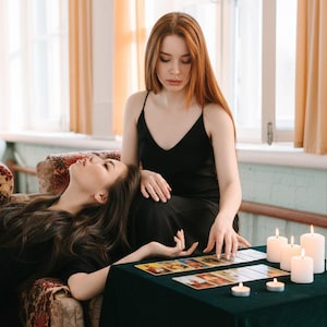 May include: Two women in black dresses are seated near a table covered with a dark green cloth. Tarot cards are laid out on the table, and several lit candles are nearby. One woman is reclining, while the other is seated and touching the cards.