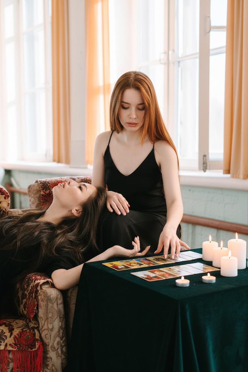 May include: Two women in black dresses are seated near a table covered with a dark green cloth. Tarot cards are laid out on the table, and several lit candles are nearby. One woman is reclining, while the other is seated and touching the cards.