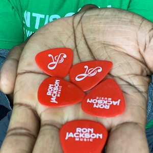 Puede incluir: Cuatro p&uacute;as de guitarra rojas con letras blancas. Tres p&uacute;as dicen "Ron Jackson Music" y una p&uacute;a tiene un s&iacute;mbolo de clave de sol.