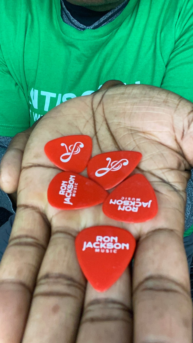 Puede incluir: Cuatro p&uacute;as de guitarra rojas con letras blancas. Tres p&uacute;as dicen "Ron Jackson Music" y una p&uacute;a tiene un s&iacute;mbolo de clave de sol.