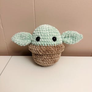 May include: A crocheted plush toy in the shape of a character with large ears. The head is light green, the body is brown, and the eyes are black. The toy is sitting on a white surface.