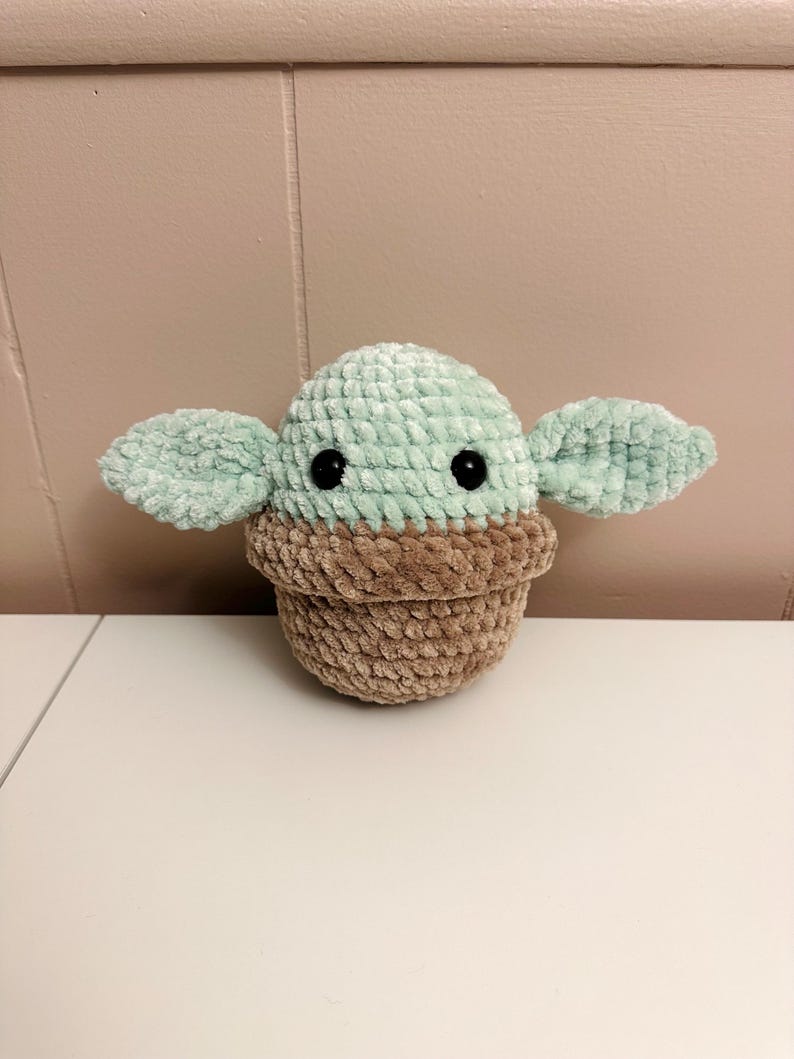 May include: A crocheted plush toy in the shape of a character with large ears. The head is light green, the body is brown, and the eyes are black. The toy is sitting on a white surface.