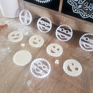 Peut inclure: Un ensemble d'emporte-pièces en plastique blanc de différentes formes, notamment des visages souriants, des cœurs et des rayures. Les emporte-pièces sont disposés sur une surface en bois légèrement saupoudrée de farine.