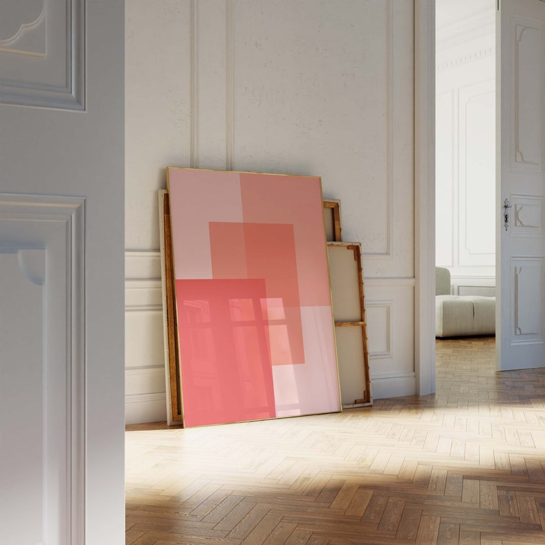 May include: A large framed artwork with a geometric design in shades of pink and coral leans against a white wall. The artwork is displayed in a room with a wooden floor and a glimpse of another room through an open doorway.
