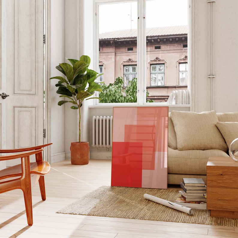 May include: A modern living room scene featuring a framed abstract art piece with red and pink geometric shapes. A large potted plant, a wooden chair, and a beige sofa are also visible. The room has a neutral color palette with natural light.