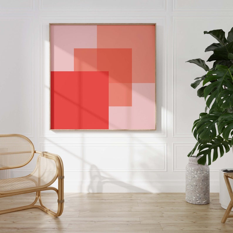 May include: A framed abstract art print featuring overlapping squares in shades of red and pink. The artwork is displayed on a white wall, with a natural wood frame. The room includes a rattan chair, a large potted plant, and a decorative vase.