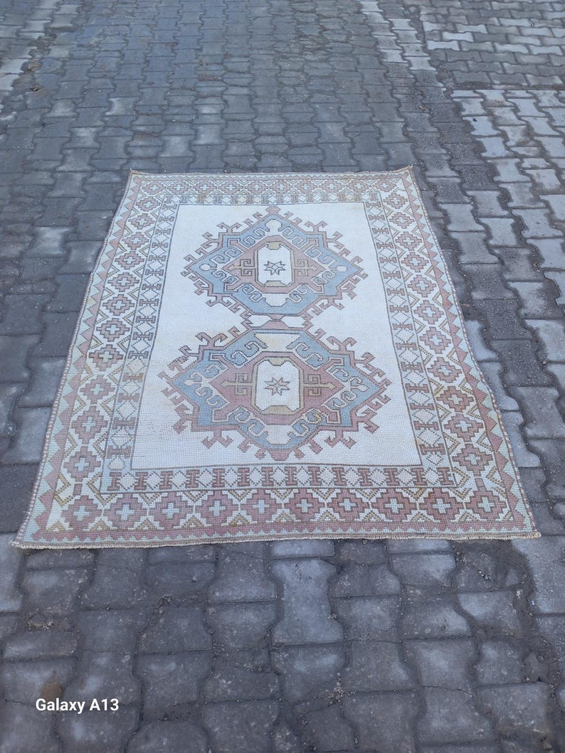 May include: A vintage rug with a cream background and a geometric pattern in shades of blue, pink, and brown. The rug has a fringed edge.