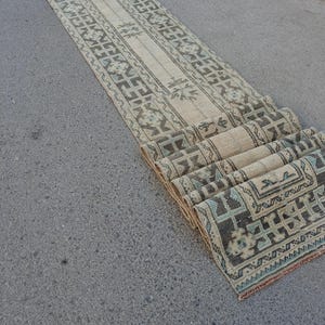 Op de afbeelding: Een lang, smal loper-tapijt met een crèmekleurige basis en ingewikkelde geometrische patronen in tinten antracietgrijs en gedempt blauwgroen. Het tapijt is aan één kant gedeeltelijk opgerold, wat een herhalend randontwerp en bloemmotieven onthult.