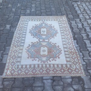 May include: A white rug with a geometric pattern in shades of pink, blue, and brown. The rug has a fringed edge and is laid out on a gray brick surface.