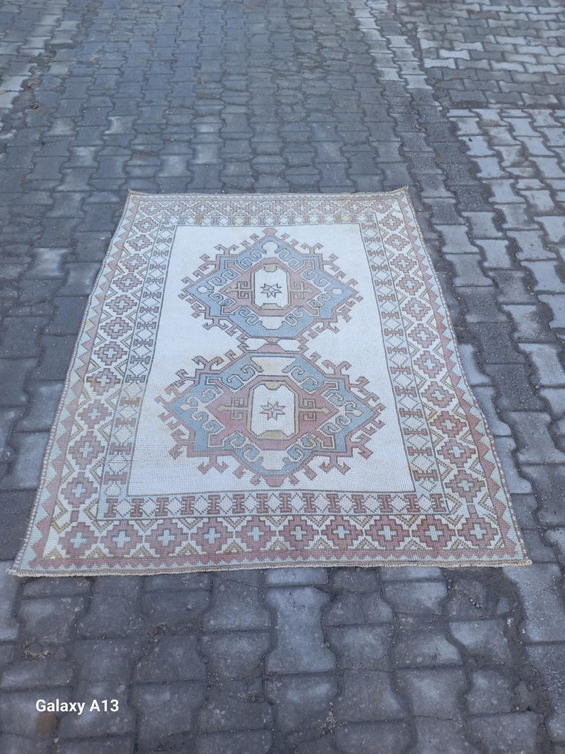 May include: A white rug with a geometric pattern in shades of pink, blue, and brown. The rug has a fringed edge and is laid out on a gray brick surface.