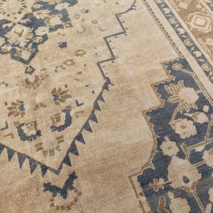 May include: Close-up of a beige and navy blue patterned rug. The rug features a central design with floral and geometric motifs. The border has a repeating pattern in shades of brown and navy blue. The rug has a distressed, vintage appearance.
