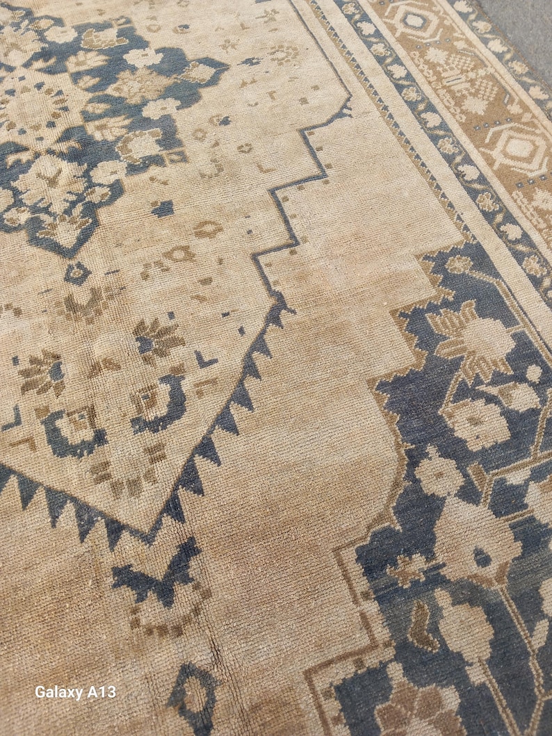 May include: Close-up of a beige and navy blue patterned rug. The rug features a central design with floral and geometric motifs. The border has a repeating pattern in shades of brown and navy blue. The rug has a distressed, vintage appearance.
