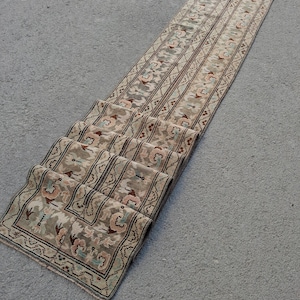 Op de afbeelding: Een lange, rechthoekige loper in beige en bruine tinten. Het tapijt heeft een herhalend patroon van bloemen en geometrische ontwerpen, met roze, blauwe en groene accenten. Het tapijt is op de voorgrond opgevouwen.