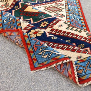 May include: A folded, rectangular rug with a detailed geometric pattern. The rug features a cream-colored central field with blue, green, red, and yellow accents. The border is red with blue and white geometric designs.