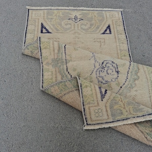May include: A small, rectangular rug with a beige and green pattern, featuring dark blue geometric shapes and borders. The rug is partially folded, revealing its underside. The rug is likely handmade and suitable for a small space.
