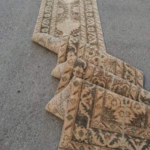 Peut inclure: Un long tapis de couloir étroit présentant un motif répétitif dans des tons beige, ocre et brun foncé. Le textile affiche des motifs géométriques et floraux complexes disposés dans un panneau central avec une bordure décorative.