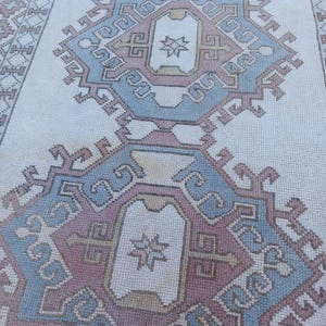 May include: A white rug with a blue and pink geometric pattern. The pattern is made up of repeating squares and diamonds. The rug has a fringe border.