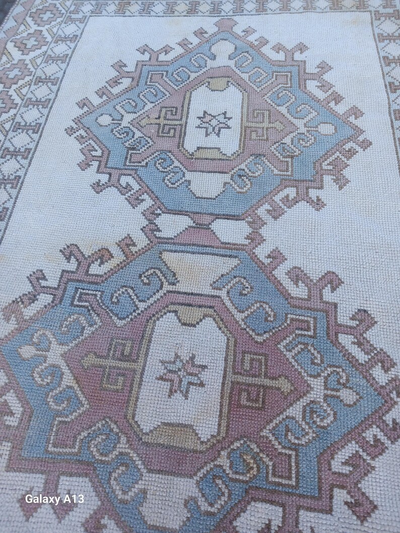 May include: A white rug with a blue and pink geometric pattern. The pattern is made up of repeating squares and diamonds. The rug has a fringe border.