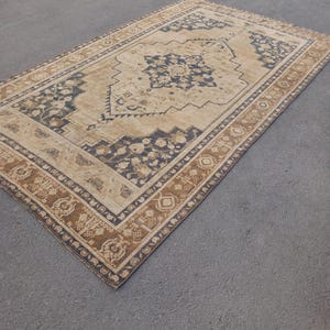 May include: A rectangular area rug with a central beige field and intricate dark blue and brown floral patterns. The rug has a detailed border with repeating motifs and a distressed, vintage appearance. The rug is placed on a gray surface.
