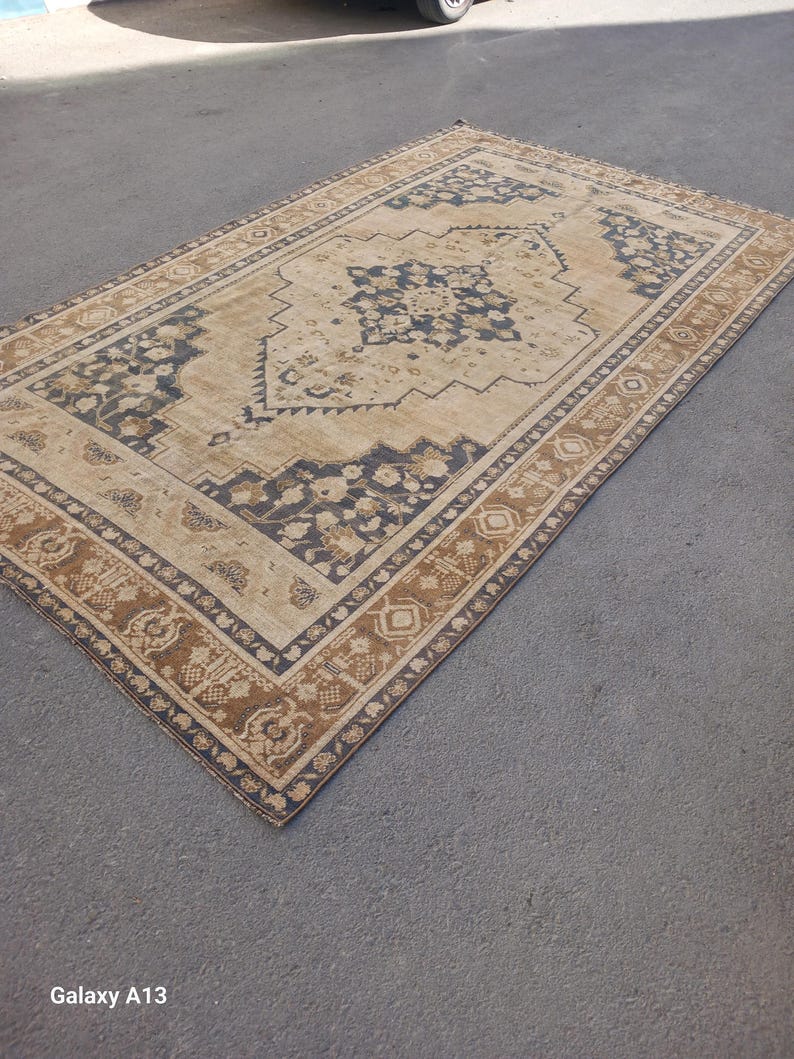 May include: A rectangular area rug with a central beige field and intricate dark blue and brown floral patterns. The rug has a detailed border with repeating motifs and a distressed, vintage appearance. The rug is placed on a gray surface.
