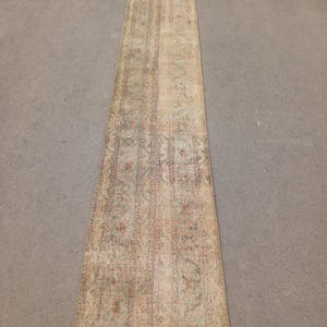May include: A long, rectangular rug with an intricate, faded floral pattern in muted tones of beige, green, and red. The rug appears to be vintage or antique, with a distressed look. The rug is placed on a gray surface.