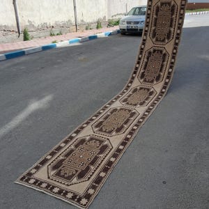 Puede incluir: Una alfombra larga y rectangular con un fondo beige y intrincados patrones geométricos marrones. La alfombra se exhibe al aire libre, mostrando su longitud y diseño detallado. Los bordes están bordeados con un ribete marrón más oscuro.