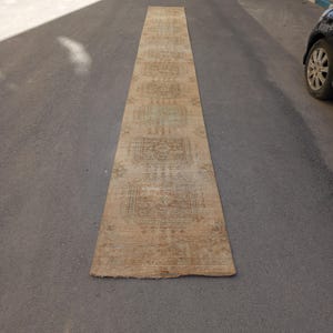 May include: A long, rectangular, beige rug with a faded, intricate pattern. The rug is laid on a grey asphalt surface, with cars parked nearby. The design features repeating geometric shapes and floral motifs.