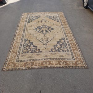 May include: Rectangular beige rug with a central navy blue medallion design and a border featuring floral and geometric patterns. The rug is placed on a gray surface, showcasing its intricate details and warm color palette.