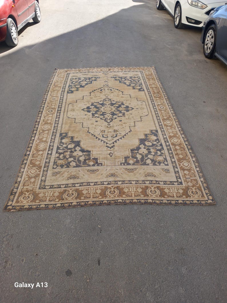 May include: Rectangular beige rug with a central navy blue medallion design and a border featuring floral and geometric patterns. The rug is placed on a gray surface, showcasing its intricate details and warm color palette.
