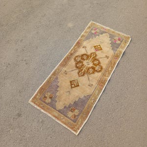 May include: A small, rectangular rug with a beige background and a brown and purple geometric pattern. The rug has a fringed border.