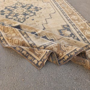 May include: A beige and brown vintage rug with a detailed floral and geometric pattern. The rug has a distressed look, with a folded corner revealing the intricate design and fringe. The rug's border is a darker brown with floral motifs.