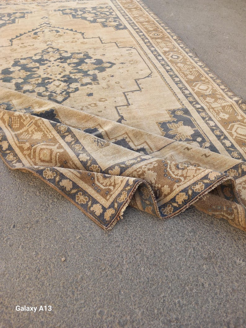 May include: A beige and brown vintage rug with a detailed floral and geometric pattern. The rug has a distressed look, with a folded corner revealing the intricate design and fringe. The rug's border is a darker brown with floral motifs.