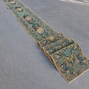 May include: A long, narrow runner rug featuring a repeating geometric and floral pattern in shades of teal, mustard yellow, and dark brown. The rug has a decorative border and is partially folded at one end, displaying its woven texture and edges.