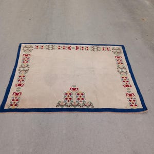 May include: A cream-colored rug with a blue border and a floral pattern in red, green, and white. The rug has a central design of a triangle with a floral pattern.