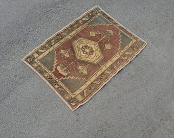 May include: A small, rectangular rug with a central geometric design in shades of red, beige, and blue. The rug features a detailed border and a faded, vintage appearance, suggesting it is handmade.
