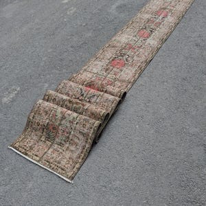May include: A long, rectangular runner rug with a faded, vintage look. The rug displays a central floral pattern in red and pink hues, set against a beige and brown backdrop. The rug is rolled up at one end, highlighting its texture and design, suitable for a hallway.