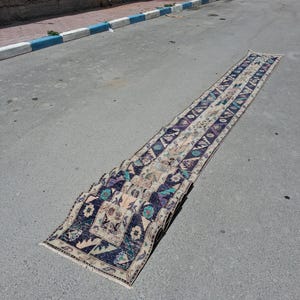 Op de afbeelding: Een lange, smalle loper met een herhalend geometrisch patroon in diep marineblauw, crème en accenten van talingblauw en gedempt roze. Het kleed heeft een rechthoekige vorm met franjes aan de uiteinden en een verweerde, versleten textuur.