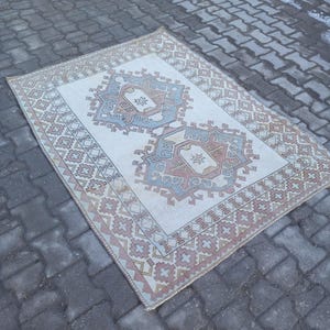 May include: A vintage cream-colored rug with a geometric pattern in shades of pink, blue, and brown. The rug has a faded, distressed look and is laid out on a gray brick surface.