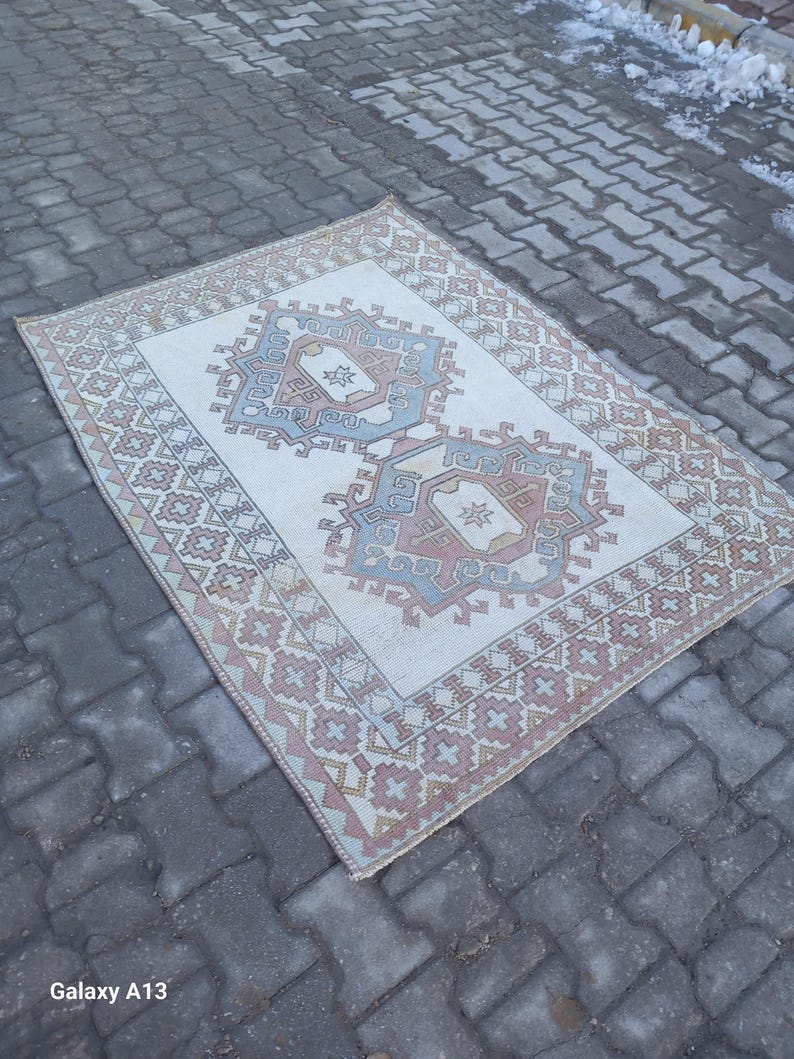 May include: A vintage cream-colored rug with a geometric pattern in shades of pink, blue, and brown. The rug has a faded, distressed look and is laid out on a gray brick surface.