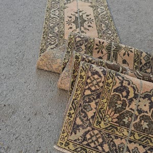 May include: A long, narrow runner rug featuring a faded peach background with intricate dark brown and olive green geometric and floral patterns. The rug has a repeating symmetrical design along its length and a short fringe at the bottom edge.