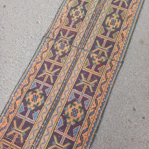 Puede incluir: Una alfombra corredora tejida con un patrón geométrico. La alfombra presenta un fondo morado oscuro con patrones repetidos en tonos melocotón, verde y azul. La alfombra es rectangular y parece estar hecha de un material duradero.