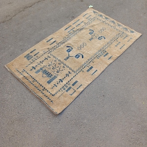 Puede incluir: Alfombra rectangular beige con un patrón geométrico azul. La alfombra presenta un diseño central con motivos florales estilizados y bordes rectangulares. La alfombra probablemente está hecha de material tejido y es adecuada para una sala de estar o dormitorio.
