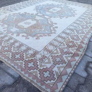 May include: A white rug with a geometric pattern in shades of pink, blue, and beige. The rug has a fringe border and is lying on a gray brick surface.