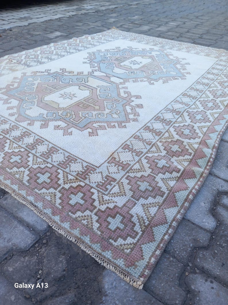 May include: A white rug with a geometric pattern in shades of pink, blue, and beige. The rug has a fringe border and is lying on a gray brick surface.