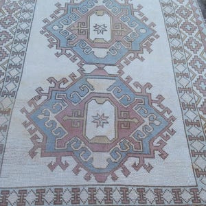 May include: A white rug with a blue, pink, and beige geometric pattern. The rug has a border with a repeating pattern of small squares and diamonds.